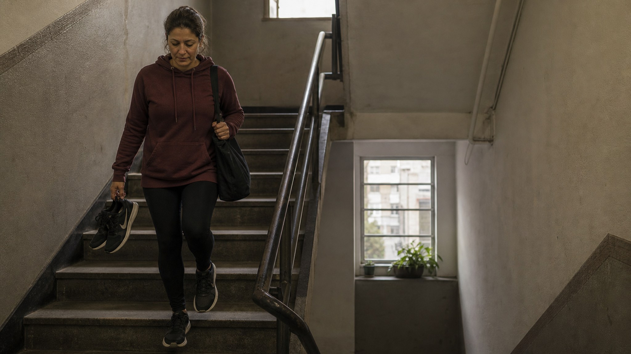 Mulher descendo a escada do pr&eacute;dio com t&ecirc;nis e bolsa de treino, mostrando a rotina antes da motiva&ccedil;&atilde;o aparecer.